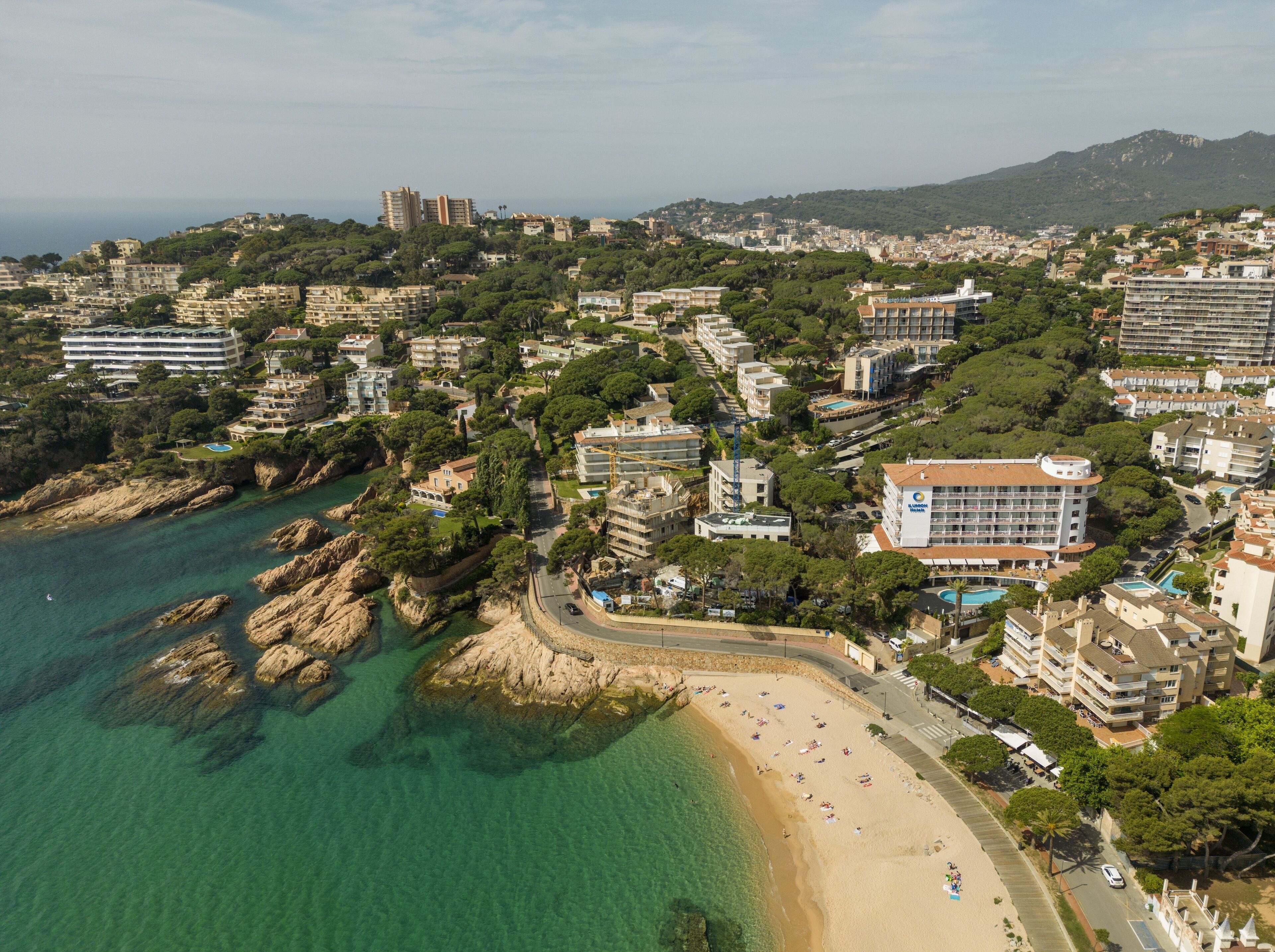 Vista aérea de un pueblo costero con playa, edificios y montañas al fondo.