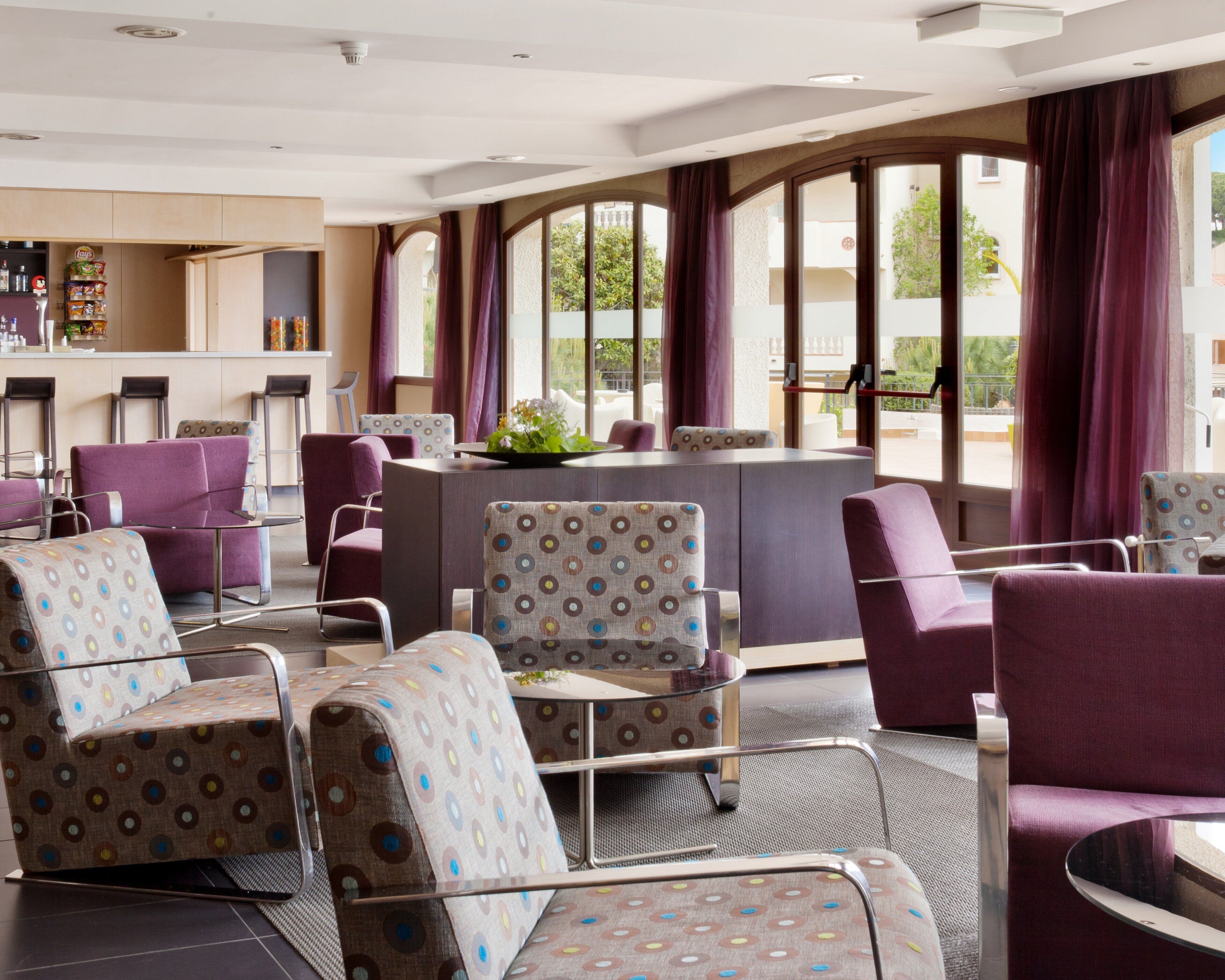 Eine Hotelhalle mit violetten und gemusterten Sesseln, einem Barbereich und großen Fenstern mit Blick ins Freie.