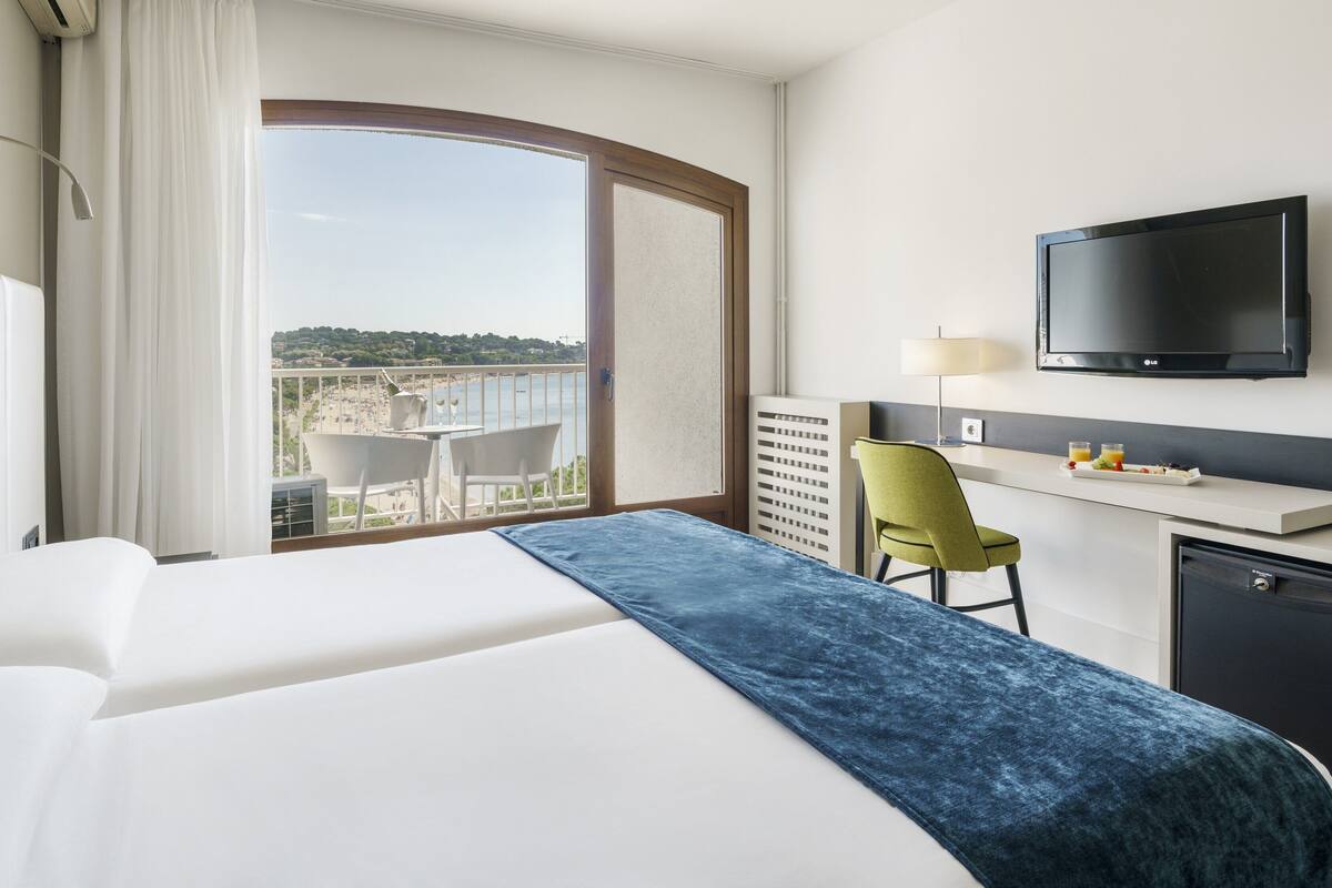 A hotel room with a bed, a desk with a chair, a television, and a view of the beach.
