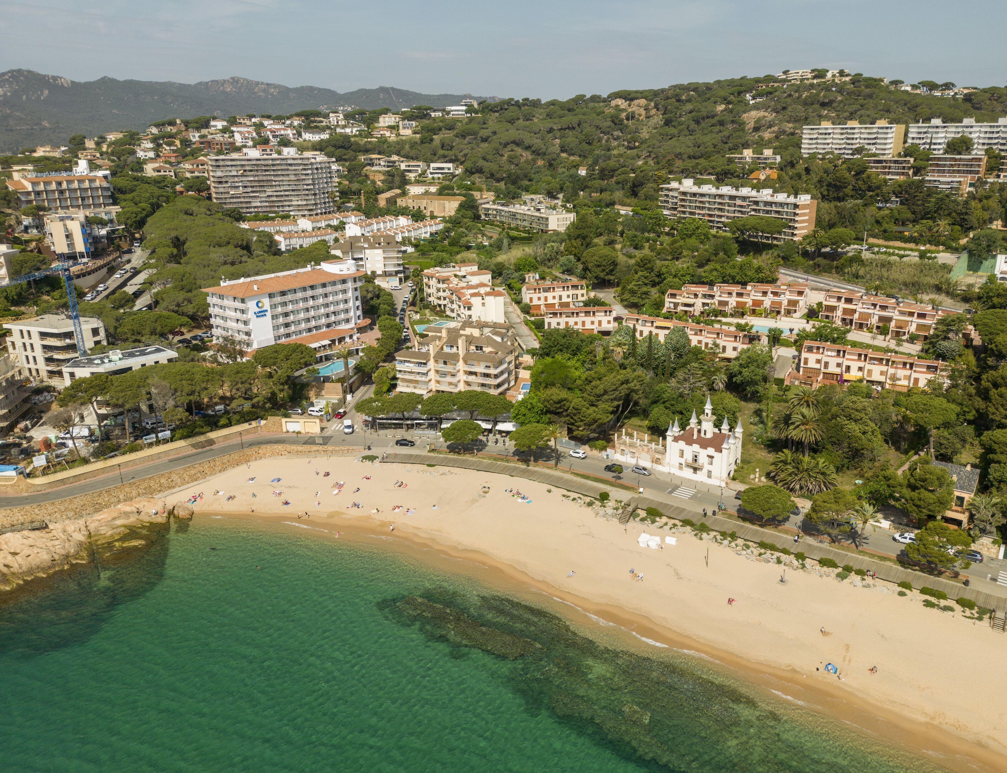 An aerial view of a coastal town with a sandy beach, a church with a bell tower, and residential buildings.