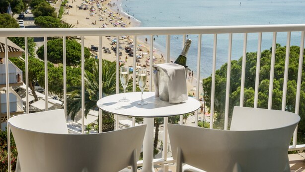 Un balcon avec deux chaises blanches et une table, donnant sur une plage où l’on peut voir des gens et des bateaux.