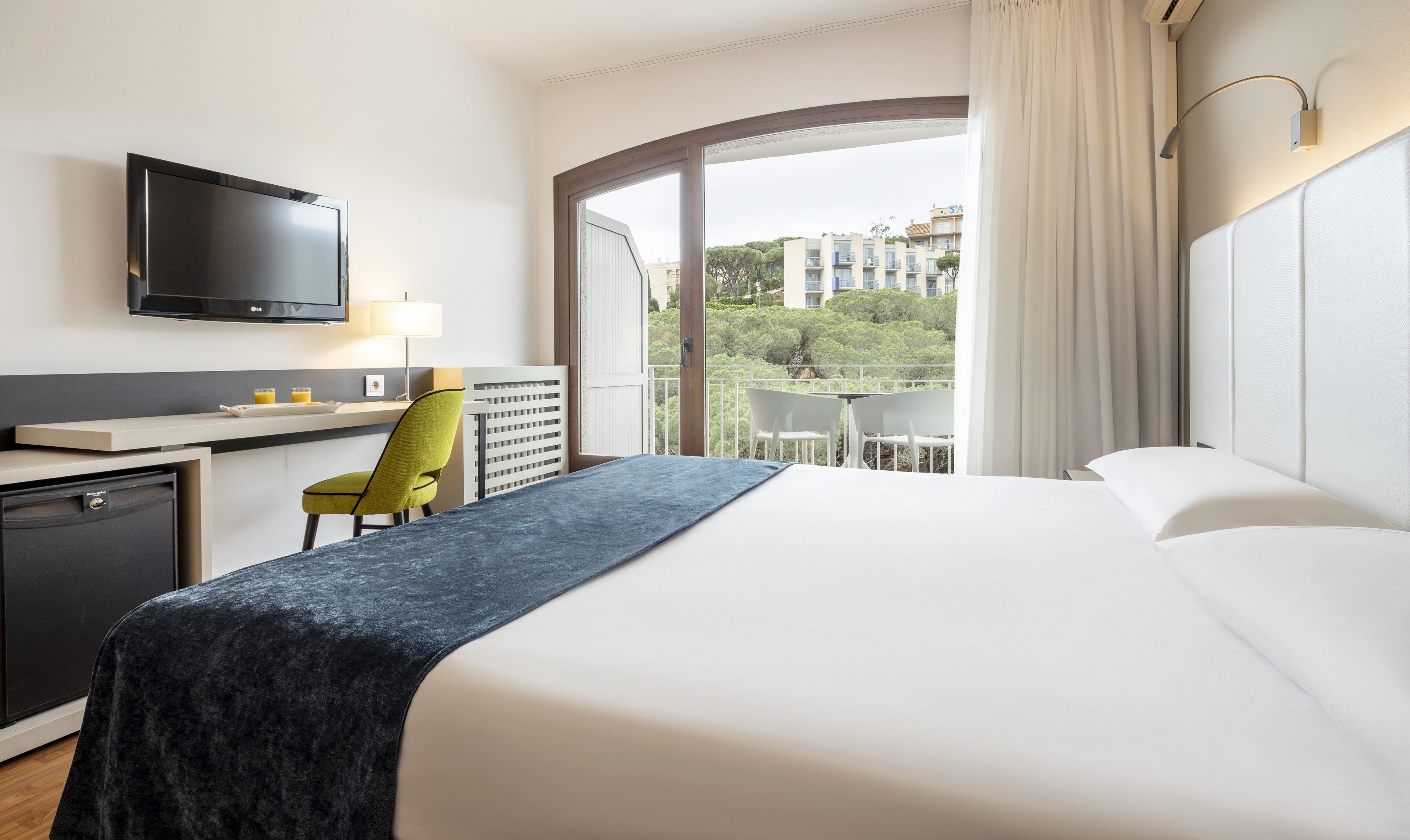 Habitación de hotel con cama, escritorio, silla, televisor y balcón con vistas a edificios y árboles.