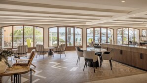 Un espace intérieur vaste et lumineux, doté de grandes fenêtres, d’un plancher en bois et de plusieurs coins salon, notamment des tables rondes et des chaises.