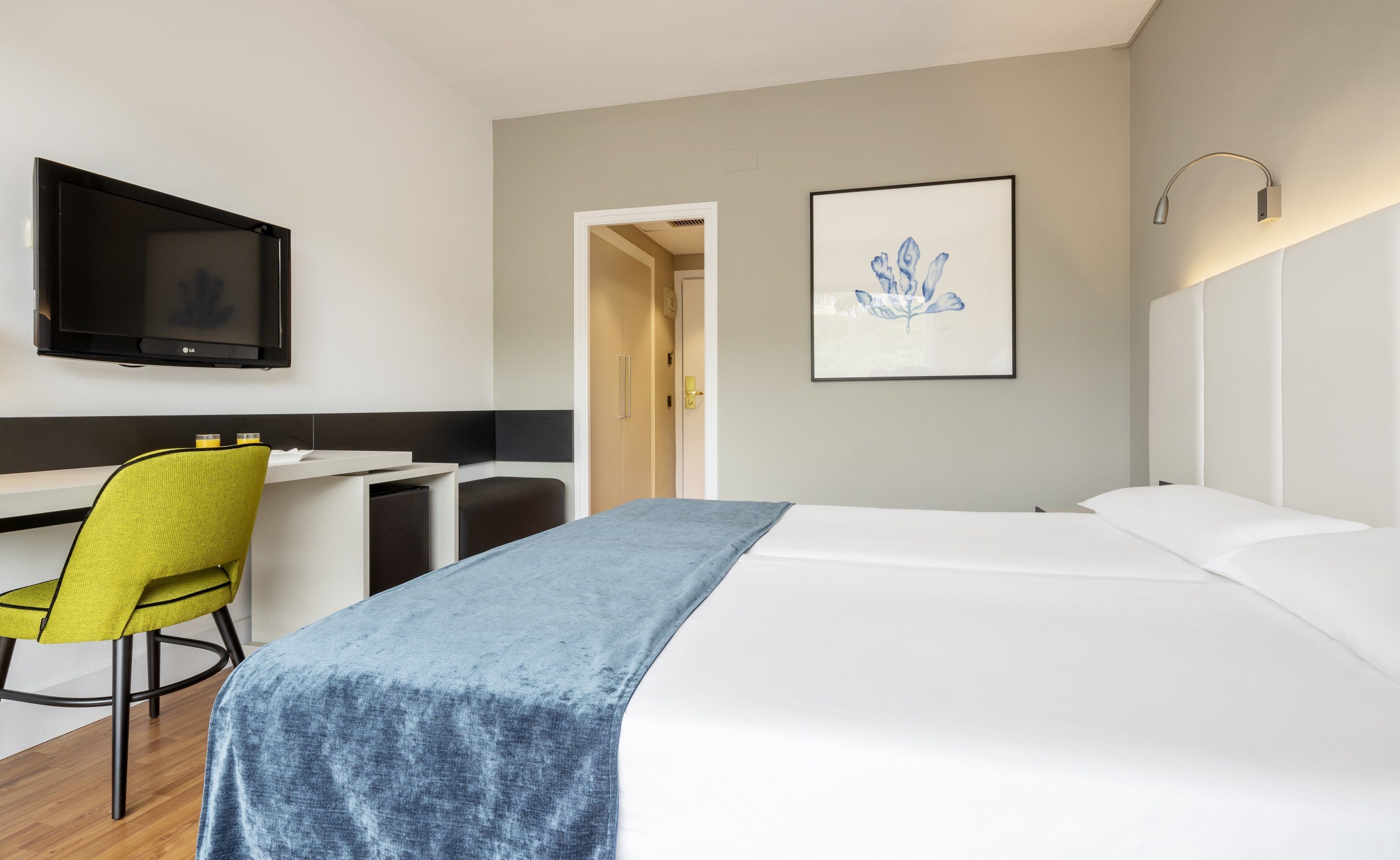 A hotel room with a bed, a desk, a chair, a TV, and a framed artwork on the wall.