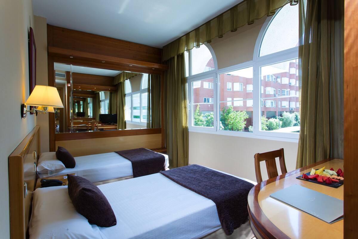 A hotel room with two beds, a dining table with fruit, a lamp, and a large window with curtains.