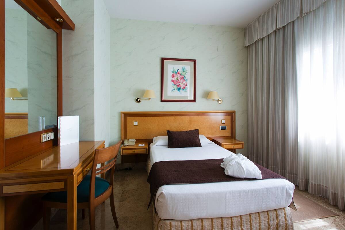 A hotel room with a bed, bedside tables, a desk, a chair, a mirror, and a framed floral artwork on the wall.