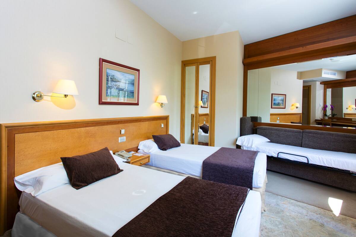 A hotel room with two beds, a sofa, a mirror, and wall-mounted lamps.