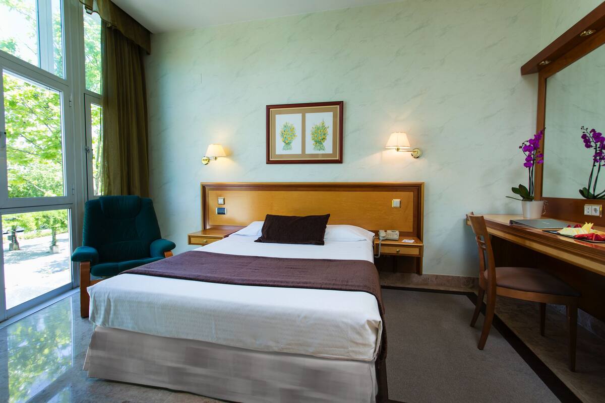 A hotel room with a bed, a desk, a chair, a nightstand, a lamp, and a window with curtains.