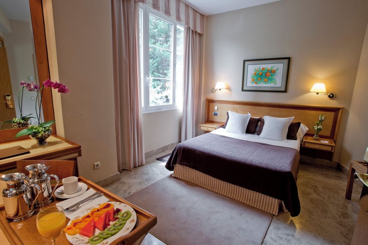A hotel room with a bed, bedside tables, a desk with a coffee maker, a fruit plate, and a window with curtains.
