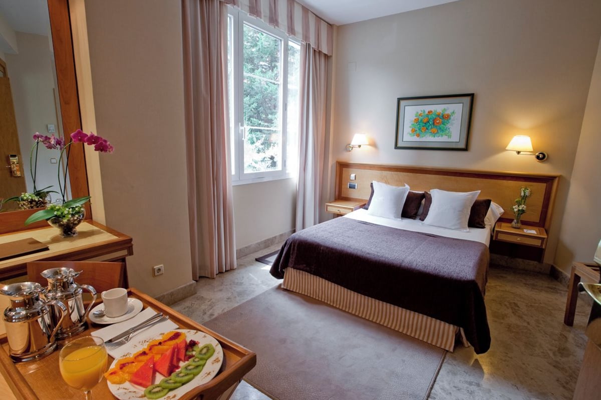 Ein Hotelzimmer mit einem Bett, Nachttischen, einem Schreibtisch mit Kaffeemaschine, einer Obstplatte und einem Fenster mit Vorhängen.