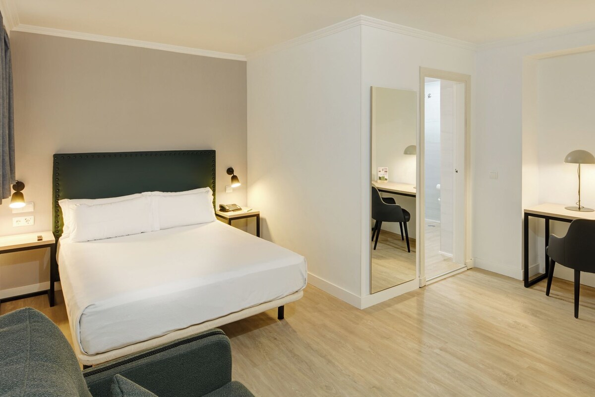 A hotel room with a bed, bedside tables, a desk, a chair, and a mirror.