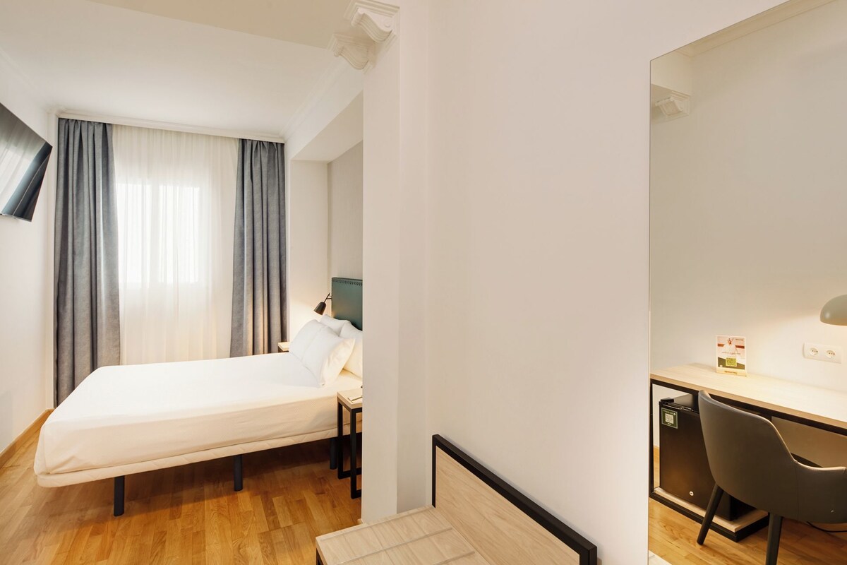 A hotel room with a bed, a desk, a chair, a TV, and a window with curtains.