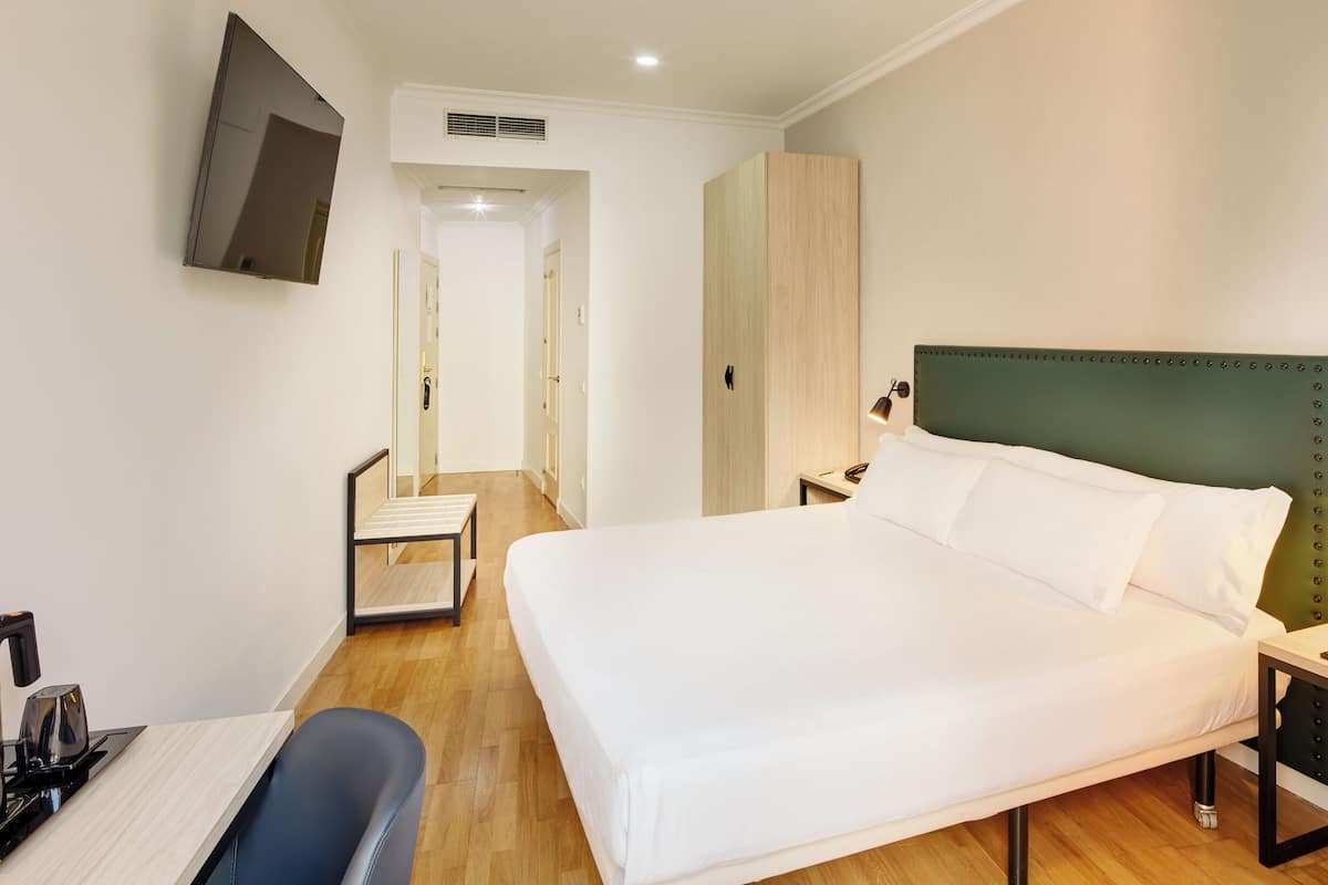 Ein Hotelzimmer mit Bett, Nachttischen, einem Schreibtisch mit Stuhl, einem an der Wand befestigten Fernseher und Holzboden.
