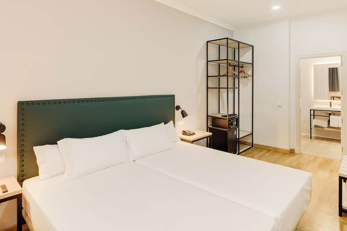 Ein ordentlich eingerichtetes Hotelzimmer mit einem großen Bett, Nachttischen, einem Kleiderschrank und einem durchgehenden Türspalt sichtbaren Badezimmer.
