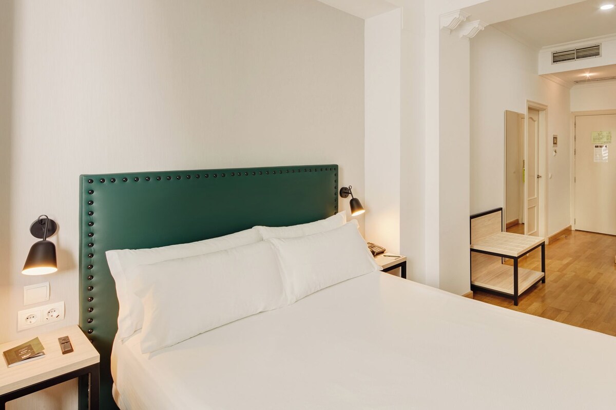 A hotel room with a green upholstered headboard, a bed with white linens, a bedside table with a lamp, a desk with a chair, and a mirror.