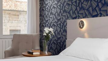 Hotelkamer met een patroonbehang, een nachtkastje met een boek en een bloevas, een bed met wit beddengoed, en een raam met uitzicht op een gebouw.