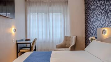 A hotel room with a bed, a desk, a chair, and a large window with sheer curtains.