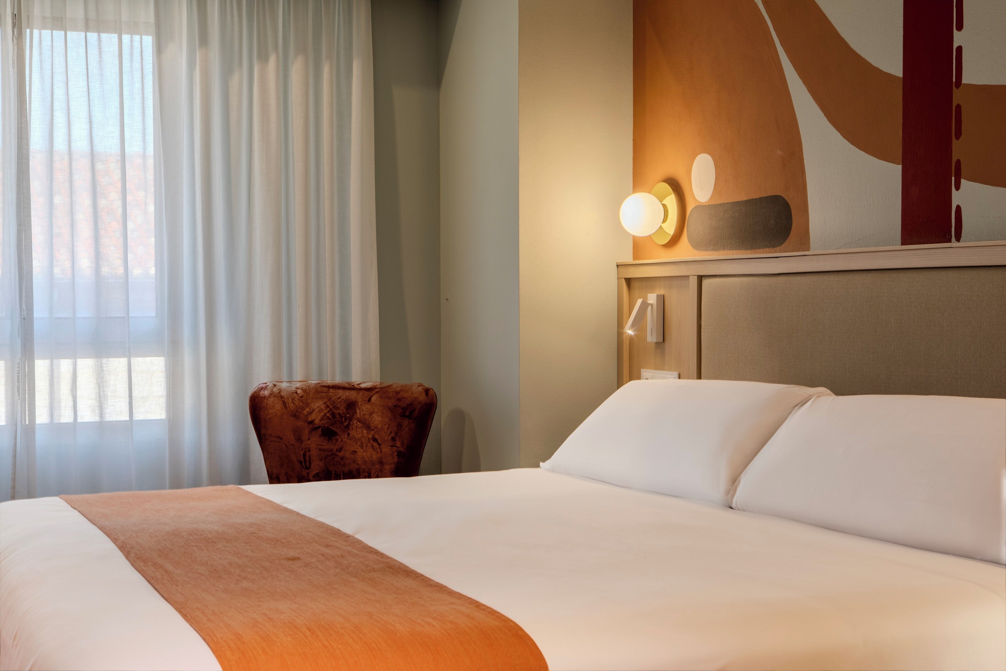 Habitación de hotel con una cama grande, una silla de madera, una lámpara de pared y un ventanal con cortinas.