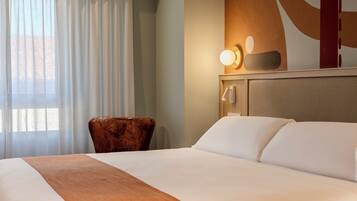 Habitación de hotel con una cama grande, una silla de madera, una lámpara de pared y un ventanal con cortinas.
