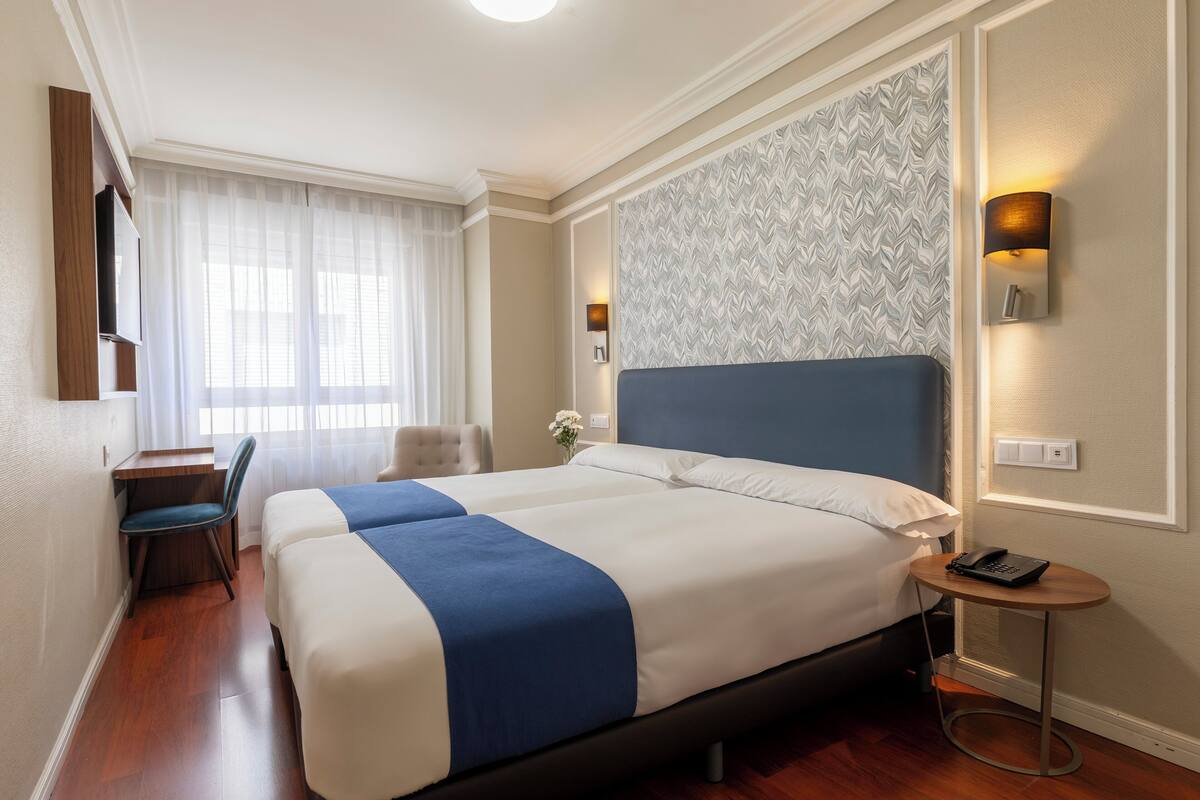 Una habitación de hotel con una cama grande, un escritorio con una silla, una mesita con un teléfono y una lámpara de pared.