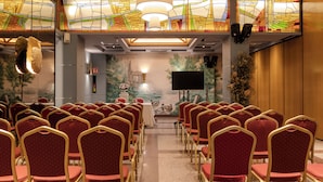 Un salón de conferencias con filas de sillas rojas y marcos dorados, una pantalla grande y un mural de un paisaje.