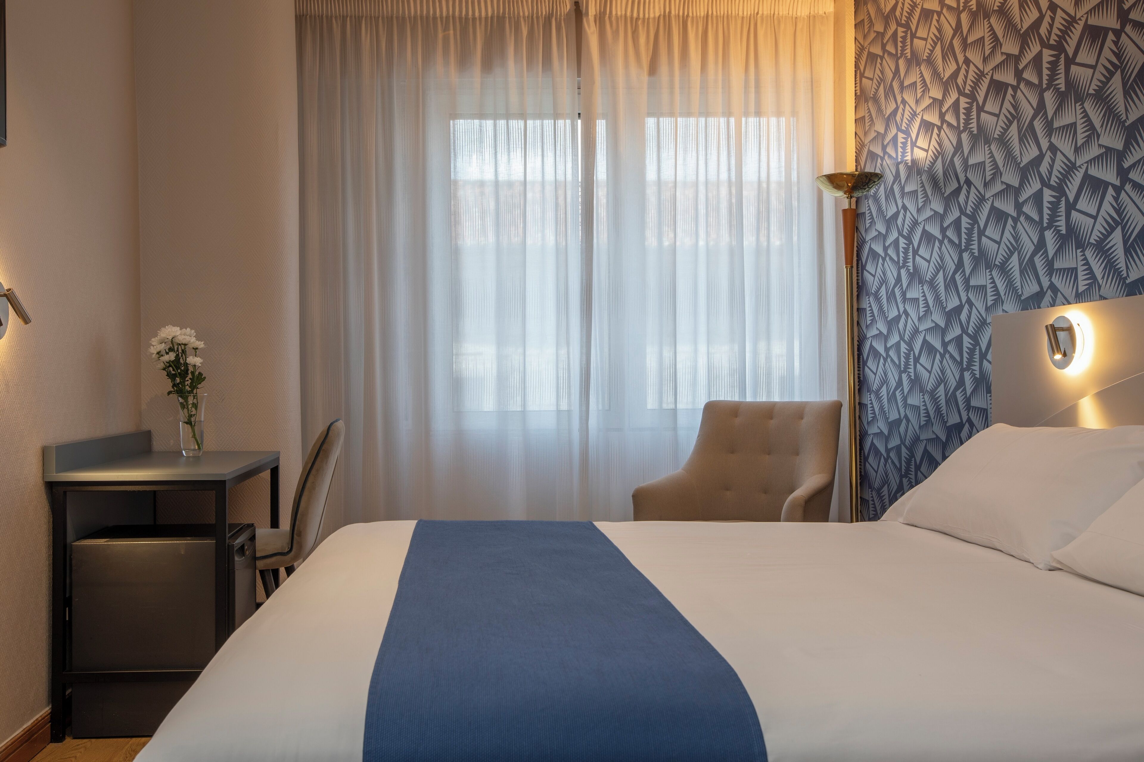 Ein Hotelzimmer mit einem großen Bett, einem Schreibtisch mit Stuhl, einem kleinen Tisch mit Blumenvase und einer Wand mit Muster.