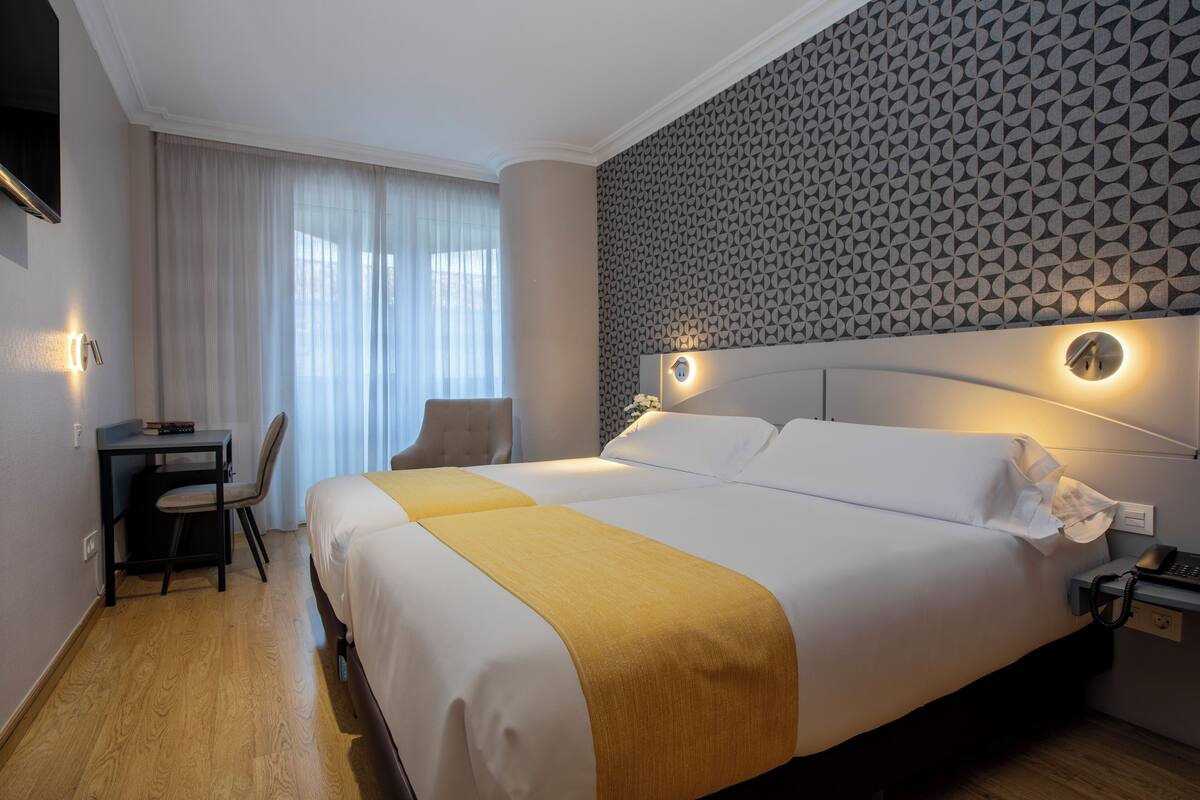 Habitación de hotel con una cama grande, un escritorio y una silla. Paredes con un diseño y una ventana con cortinas.