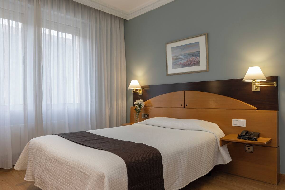 Habitación de hotel con una cama grande, dos lámparas de noche, una mesita de noche con un teléfono y un cuadro en la pared.