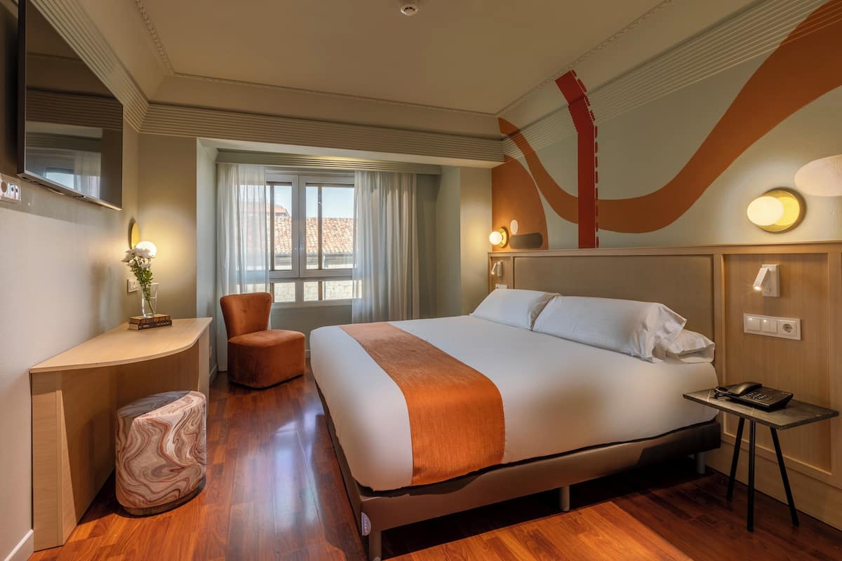 Habitación de hotel con una cama grande, un escritorio, una silla, un pequeño reposapiés, una mesita de noche con teléfono y una ventana con cortinas.