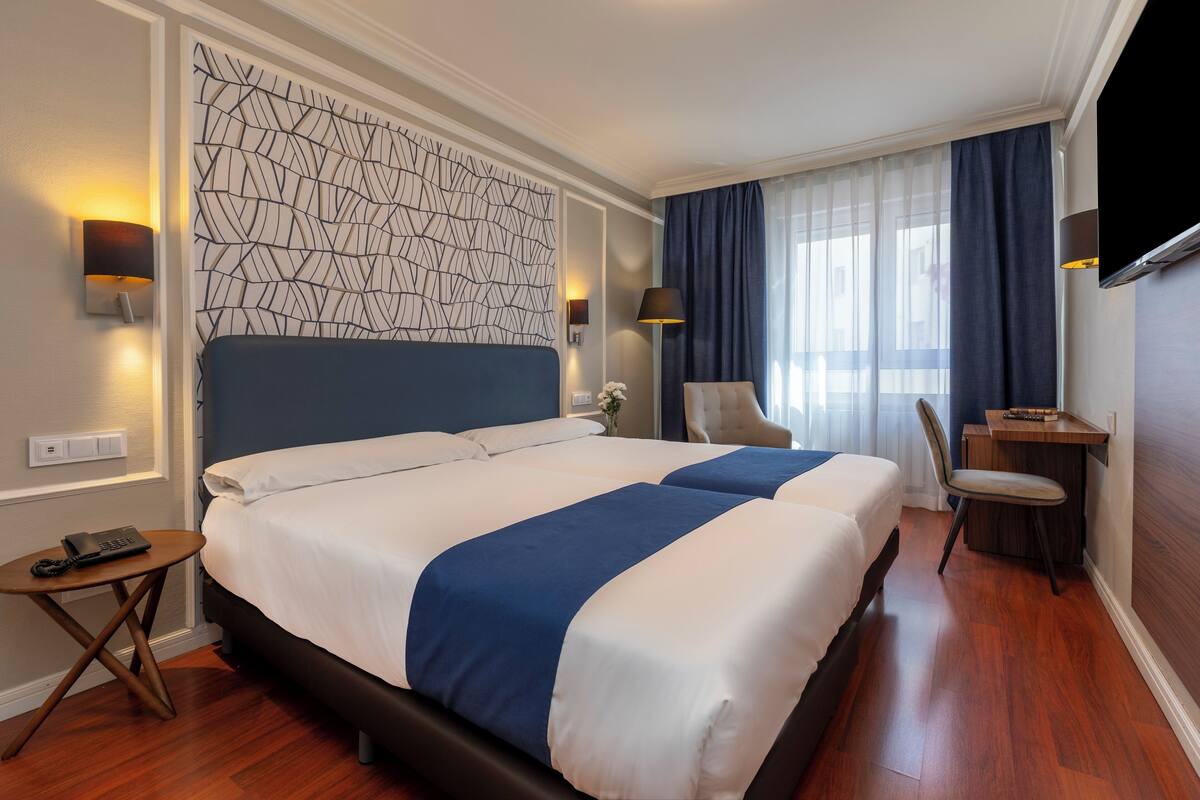 Habitación de hotel con dos camas, un escritorio, una silla y un televisor.