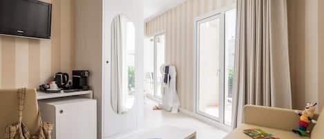 A hotel room with a flat-screen TV, a coffee machine, a sofa, a child’s play area, and a large window with curtains.