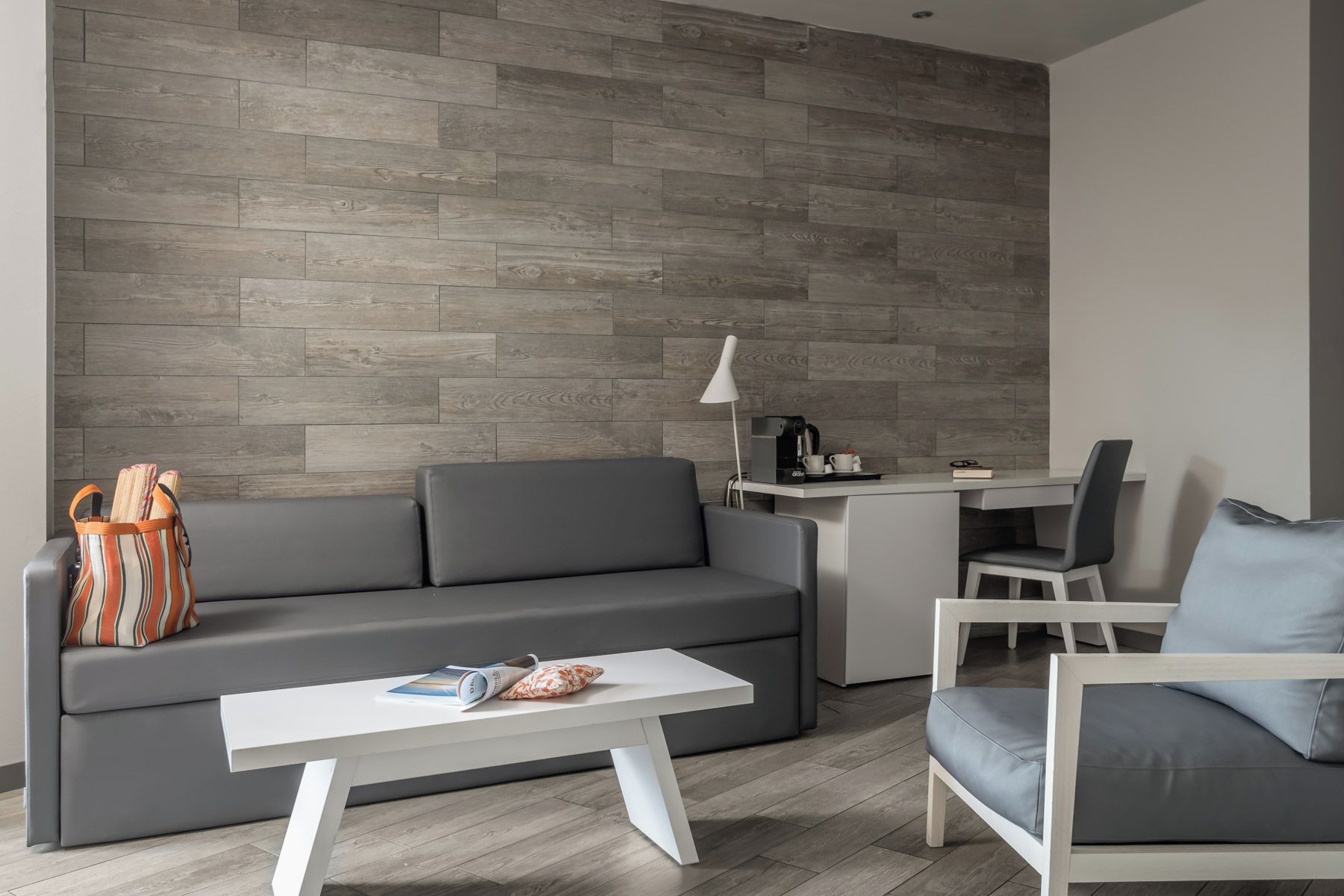 Una sala de estar moderna con un sofá gris, una mesa de centro blanca, un bolso a rayas y un escritorio con una lámpara.