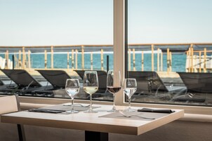 Uma mesa com quatro copos de vinho, um smartphone e um menu em um restaurante à beira-mar, com vista para o oceano e um píer de madeira.