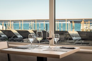 Una mesa con cuatro copas de vino, un teléfono inteligente y un menú en un restaurante frente al mar, con vista al océano y un muelle de madera.