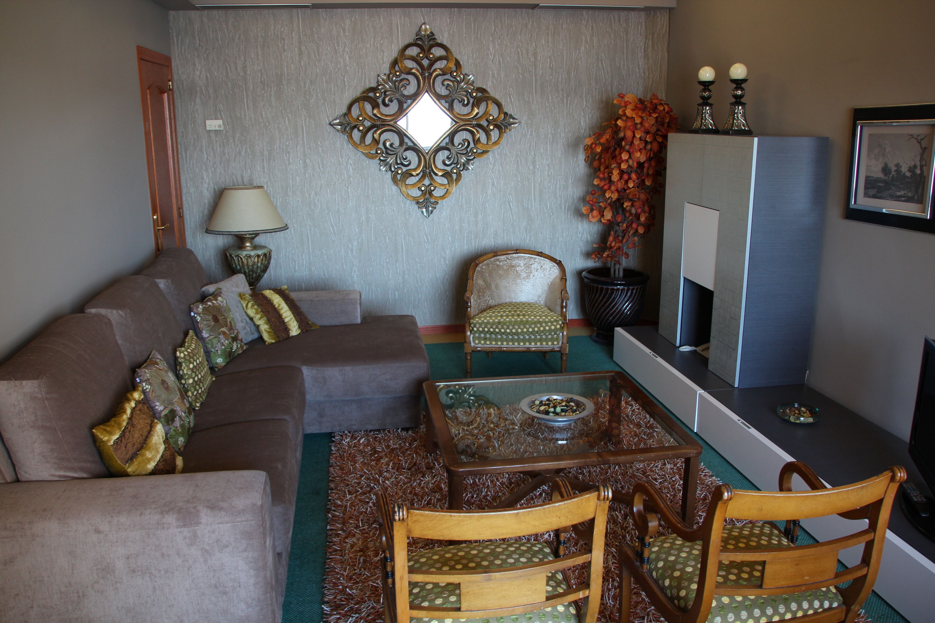 A living room with a sofa, two chairs, a glass-top coffee table, a decorative mirror, a lamp, a fireplace, and a television.