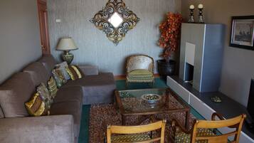 Un salon comprenant un canapé, deux fauteuils, une table basse avec un plateau en verre, un miroir décoratif, une lampe, une cheminée et un téléviseur.