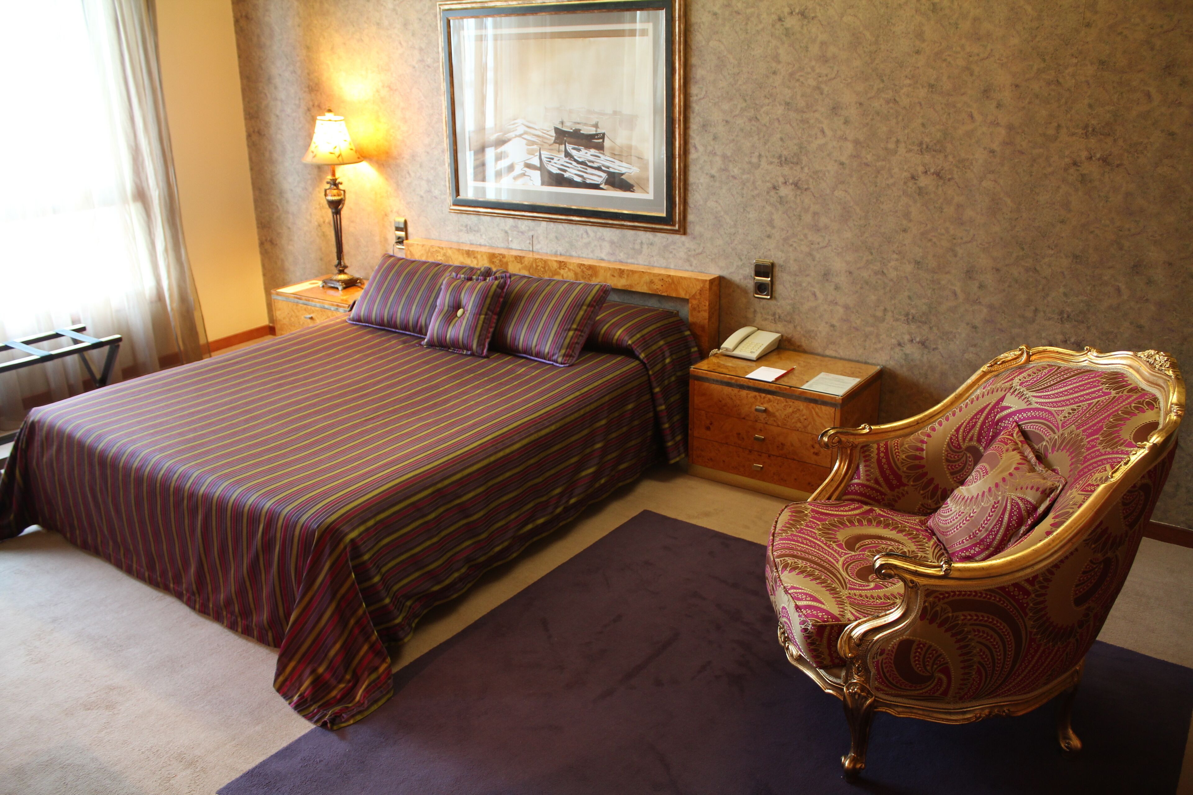 A hotel room with a bed, a chair, a nightstand, a lamp, and a framed picture on the wall.