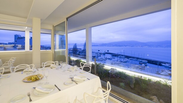 A dining area with tables set for a meal, large windows offering a view of a marina and cityscape. - Hotel Ciudad de Vigo (Vigo)