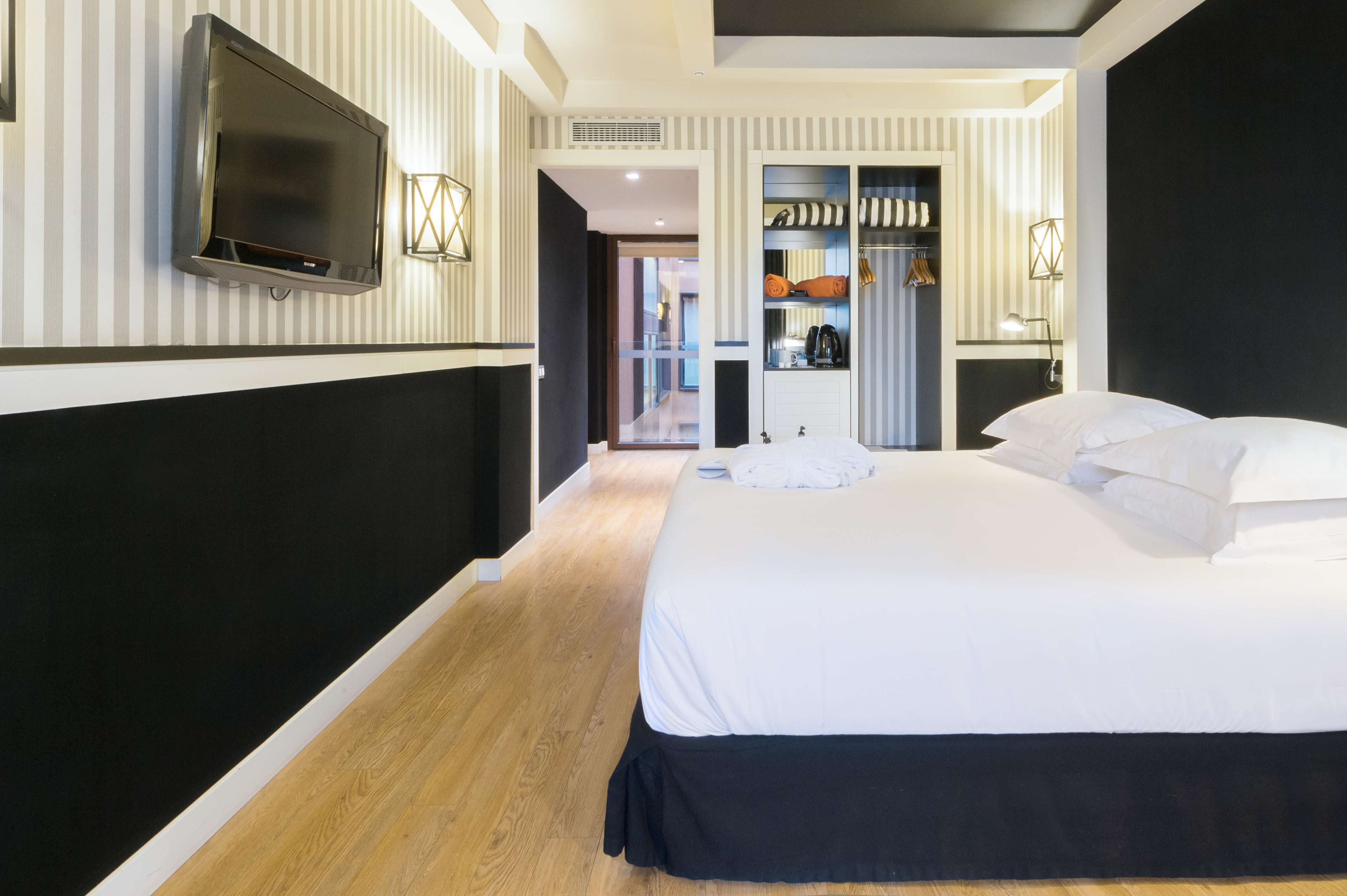 A modern hotel room with a large bed, a flat-screen TV mounted on the wall, a walk-in closet, and a hallway leading to other rooms.