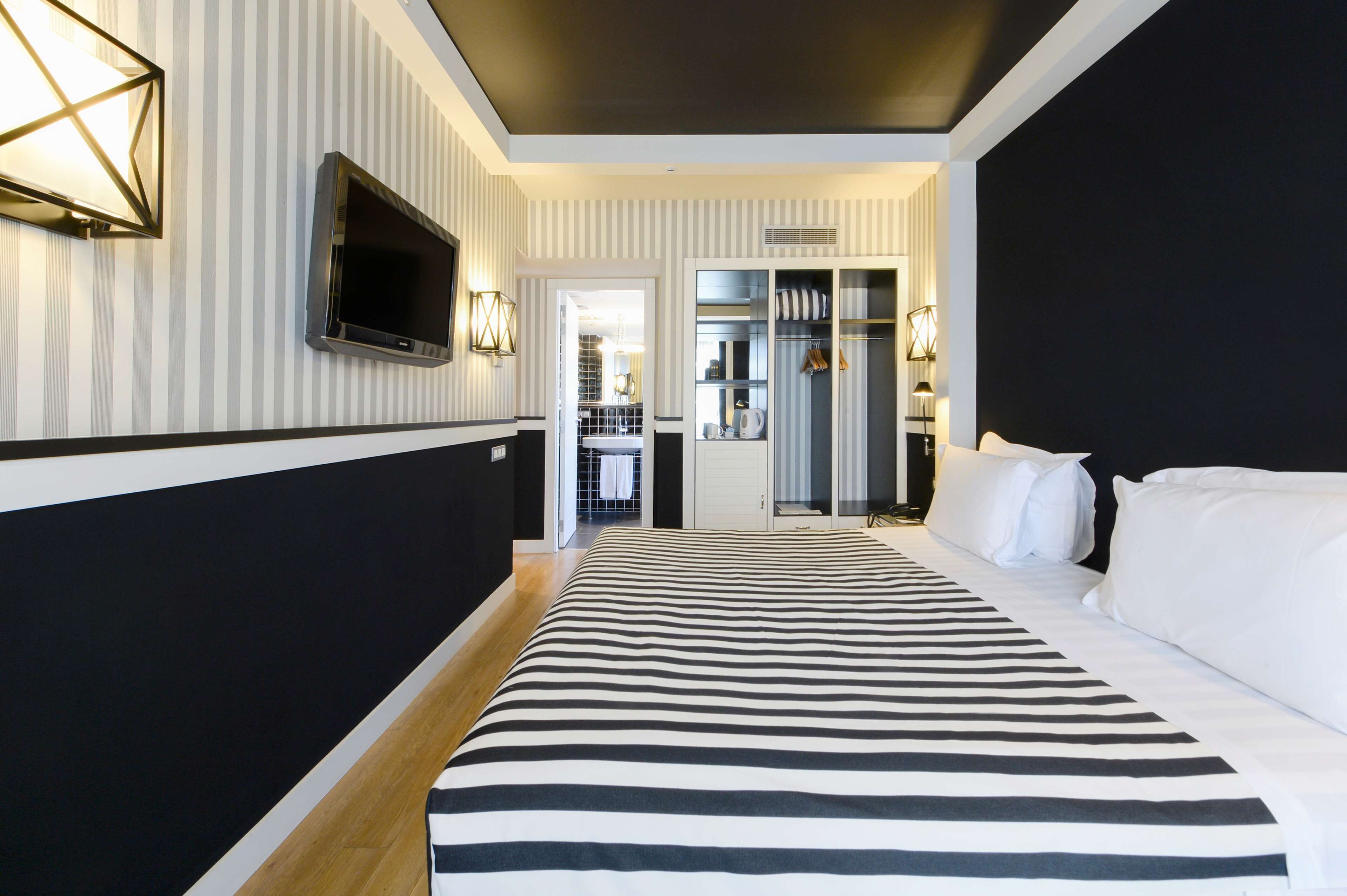 Habitación de hotel con pared a rayas, una cama grande con ropa de cama blanca y negra, un televisor de pantalla plana montado en la pared y un vestidor con espejo y ropa colgada.
