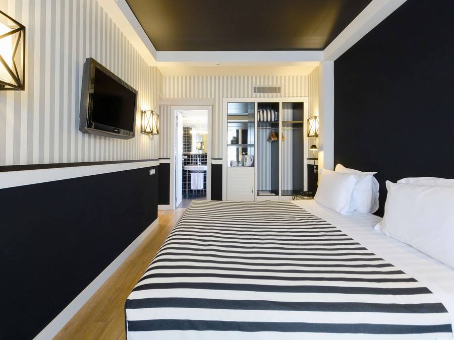 Habitación de hotel con pared a rayas, una cama grande con ropa de cama blanca y negra, un televisor de pantalla plana montado en la pared y un vestidor con espejo y ropa colgada.