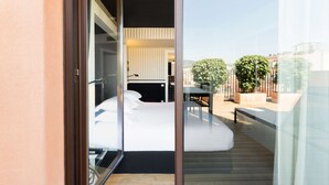 Habitación de hotel moderna con una puerta de vidrio que da a un balcón. La habitación tiene una cama, un escritorio y una silla. El balcón tiene piso de madera y está rodeado de follaje y edificios.