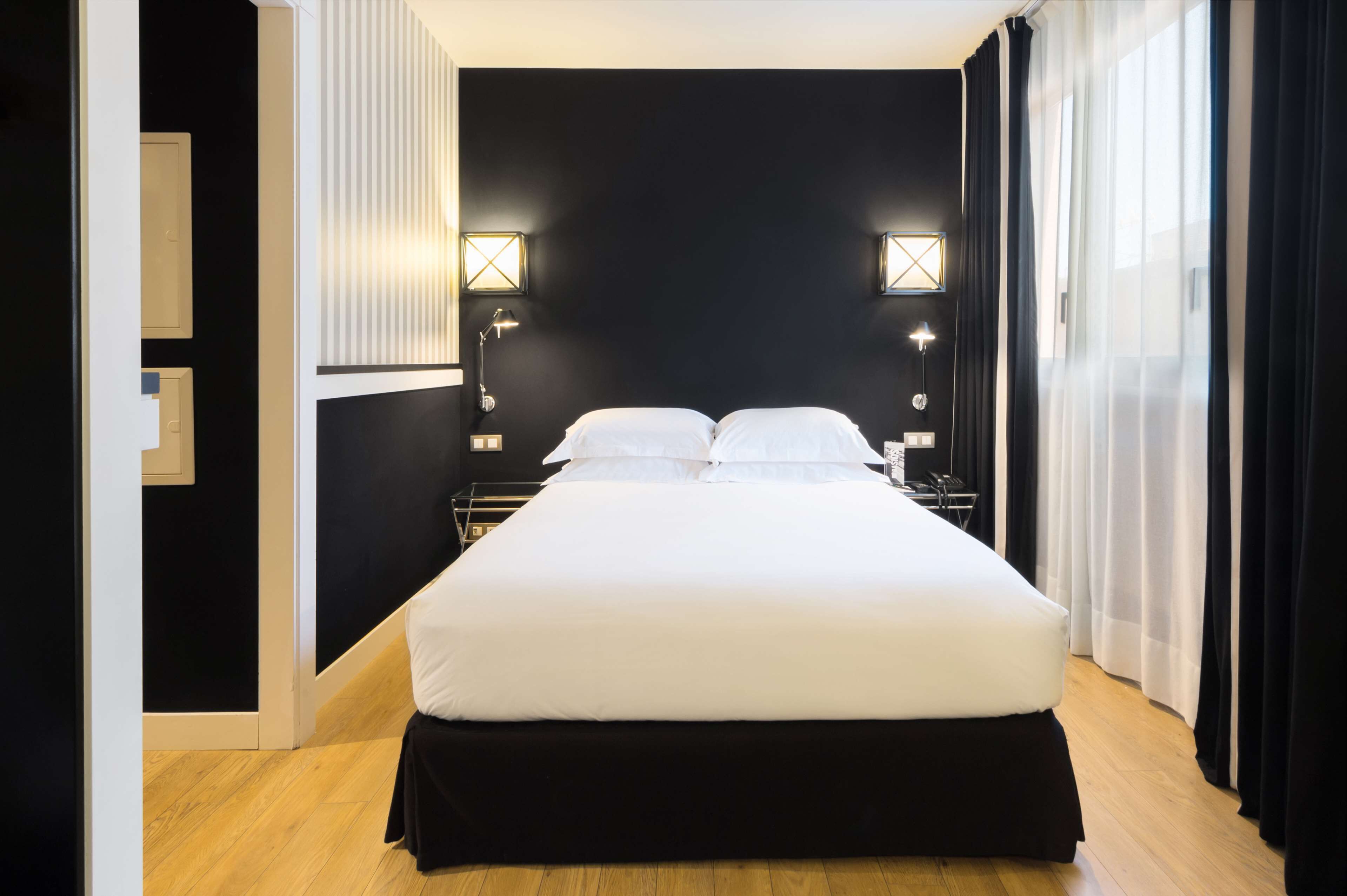 A modern hotel room with a large bed, two bedside lamps, and a view of the outdoors through sheer curtains.