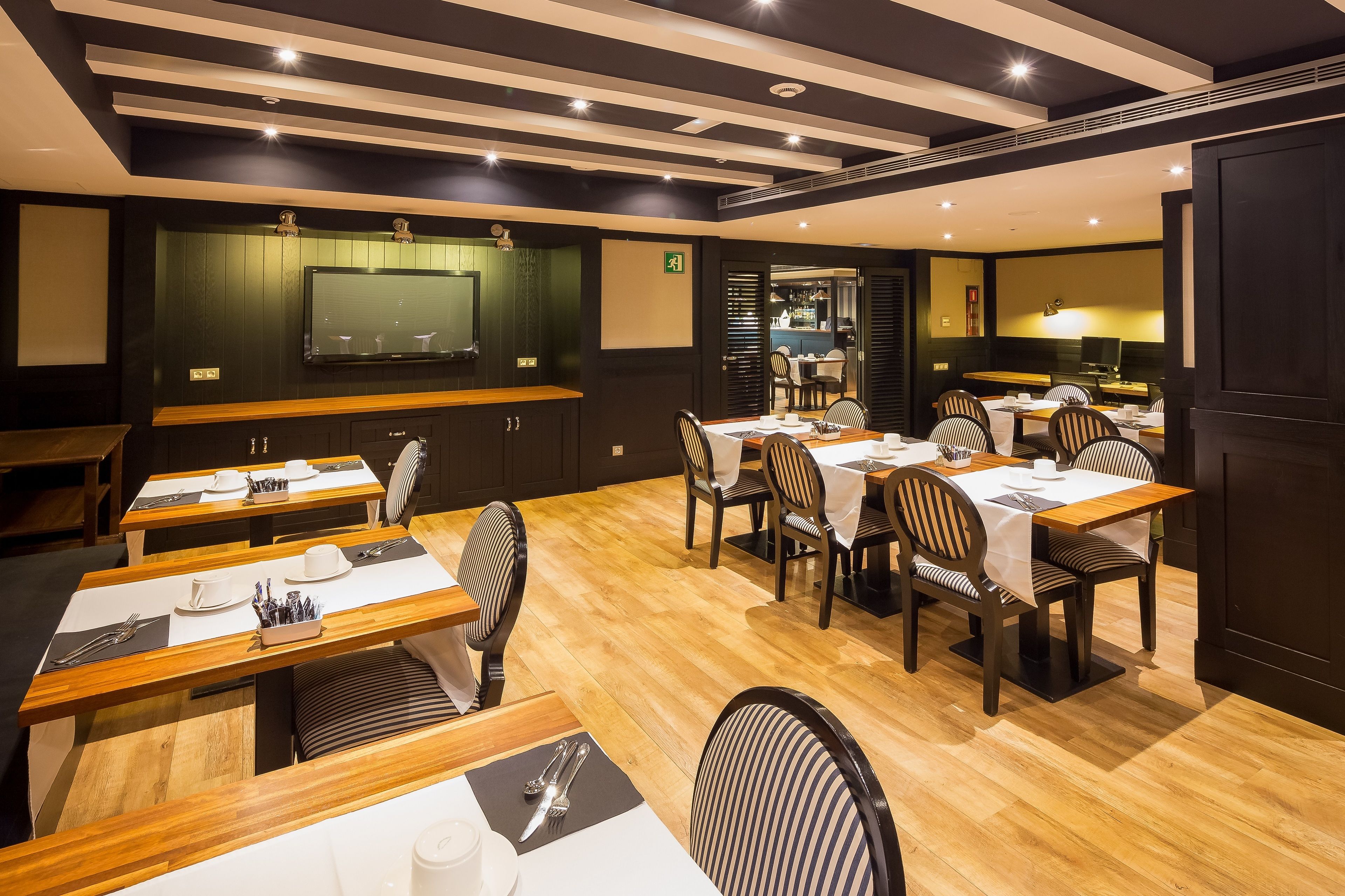 Un restaurant avec des tables et des chaises en bois, un téléviseur fixé au mur et un plafond doté d’un éclairage encastré.
