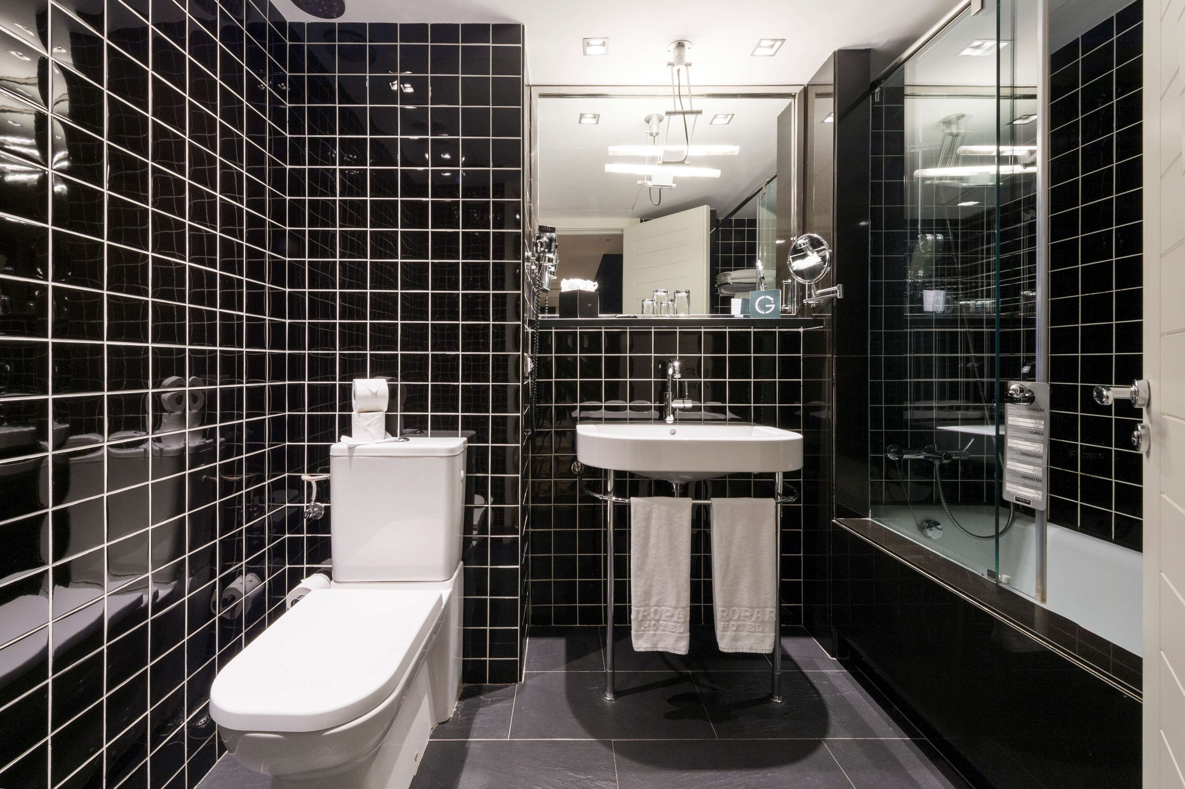 Un baño moderno con paredes de azulejos negros, un inodoro blanco, un lavabo y una zona de ducha con mampara de cristal.