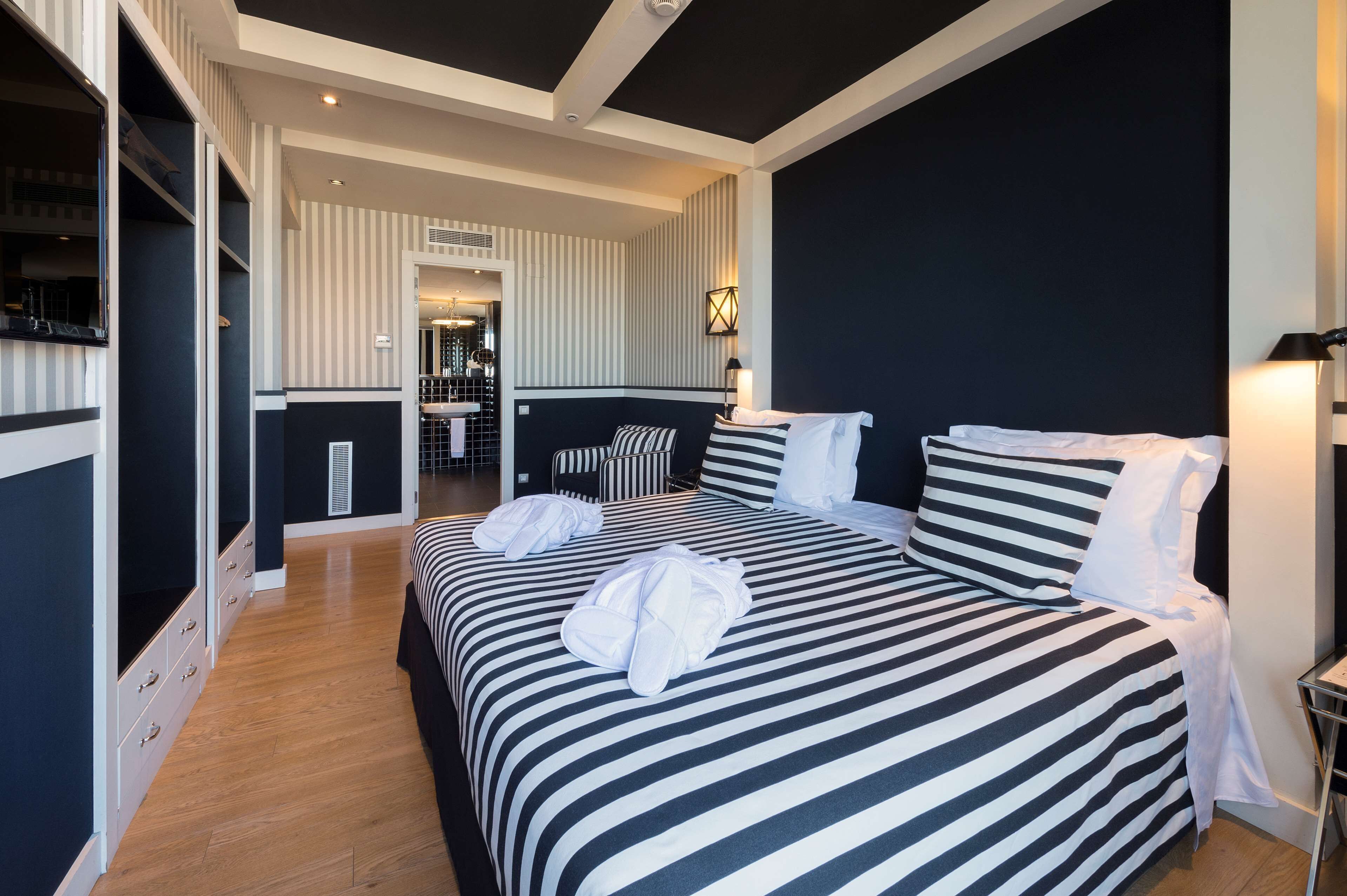 Habitación de hotel con una cama grande, ropa de cama a rayas, una mesita de noche, una lámpara y vistas al baño.