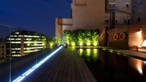 Una piscina en la azotea con cubierta de madera, rodeada de vegetación e iluminada con luces tenues.