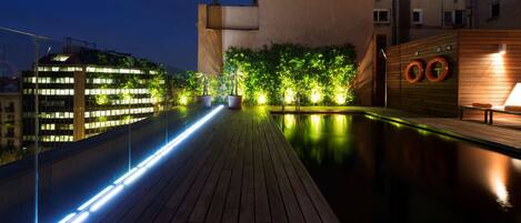 Una piscina en la azotea con cubierta de madera, rodeada de vegetación e iluminada con luces tenues.