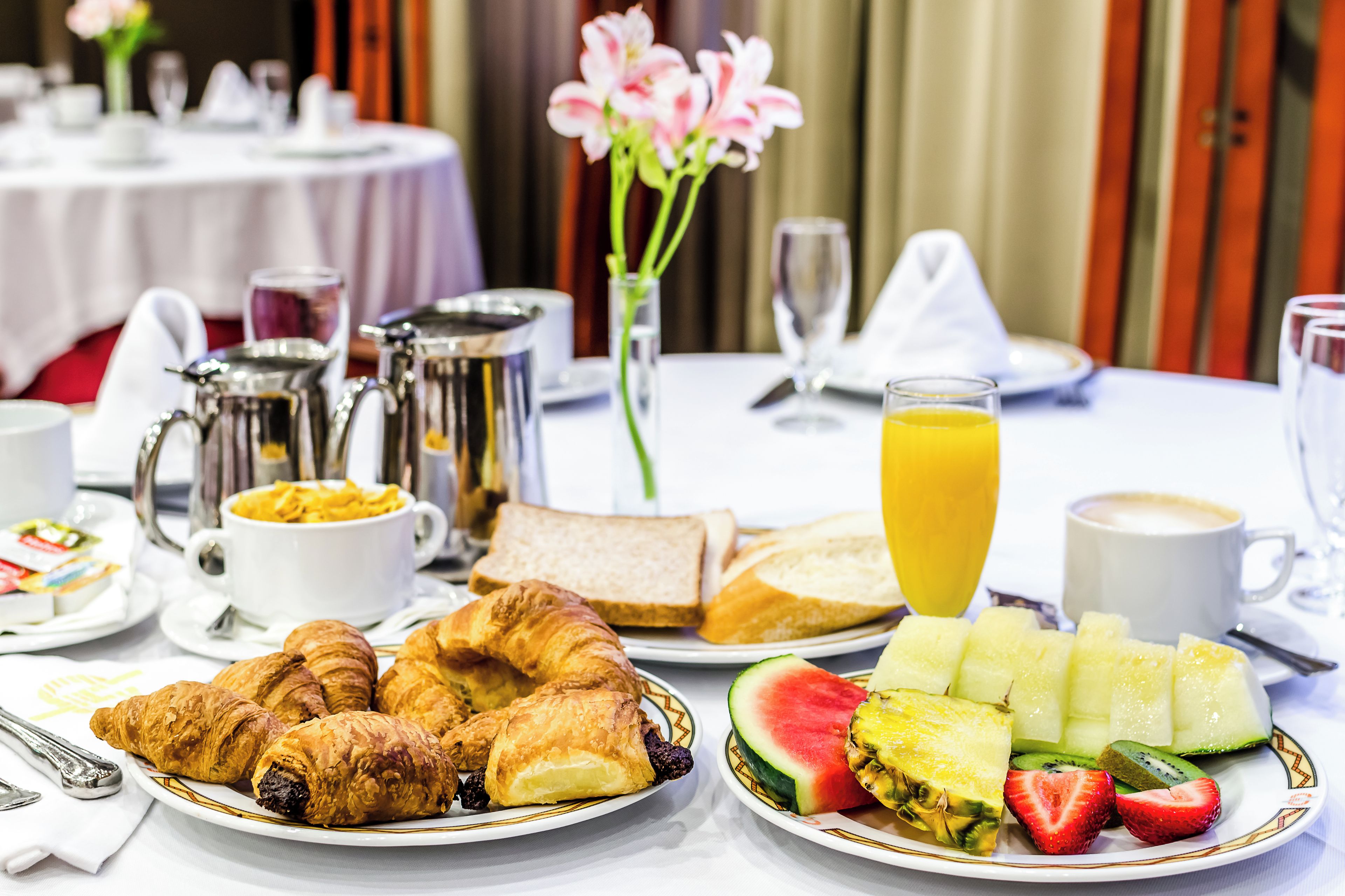 Desayuno con cruasanes, pan, fruta y bebidas sobre una mesa.