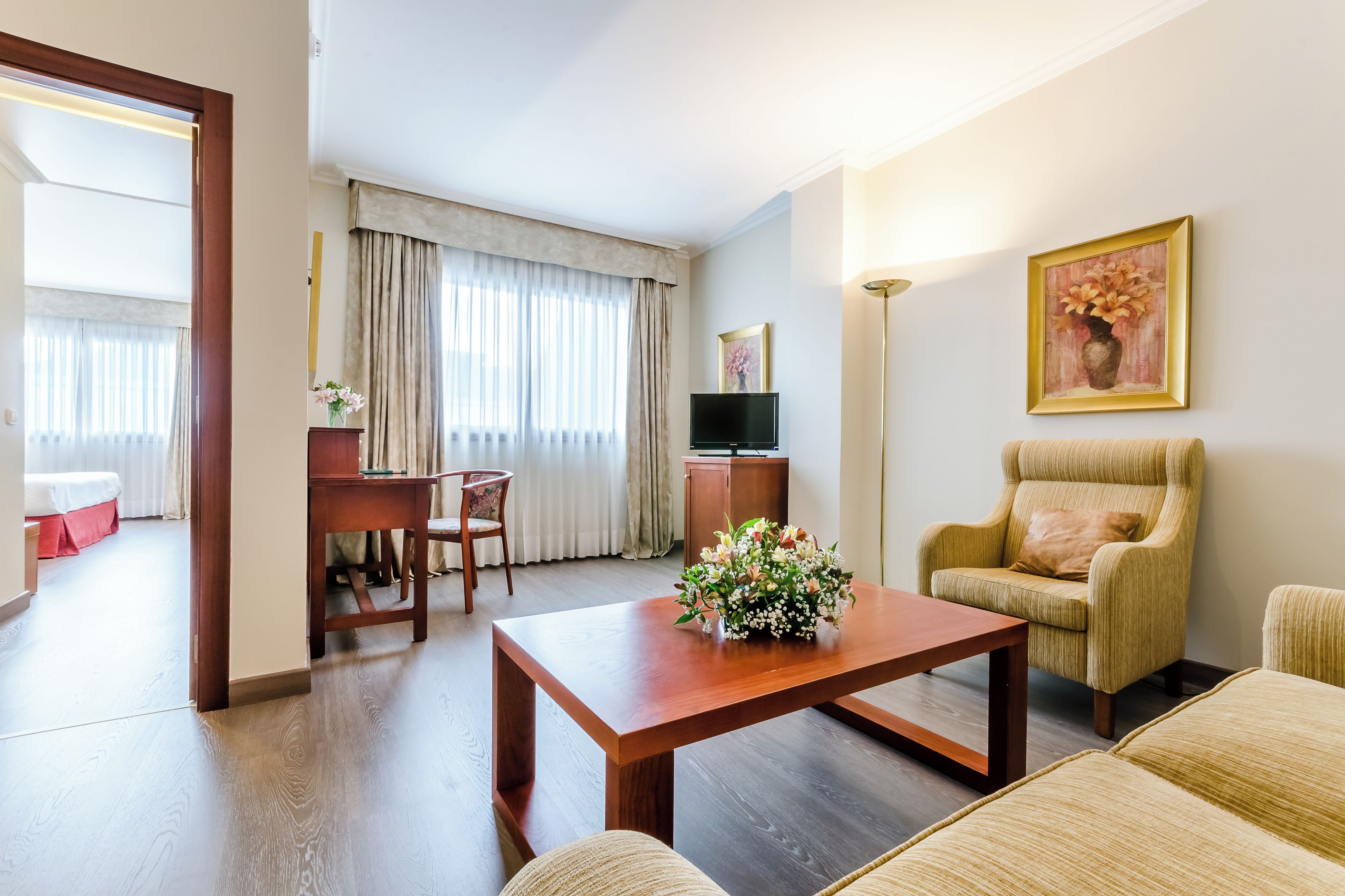 Habitación de hotel con piso de madera, un escritorio con silla, un sofá, una mesa de centro con flores, un televisor y un cuadro en la pared.